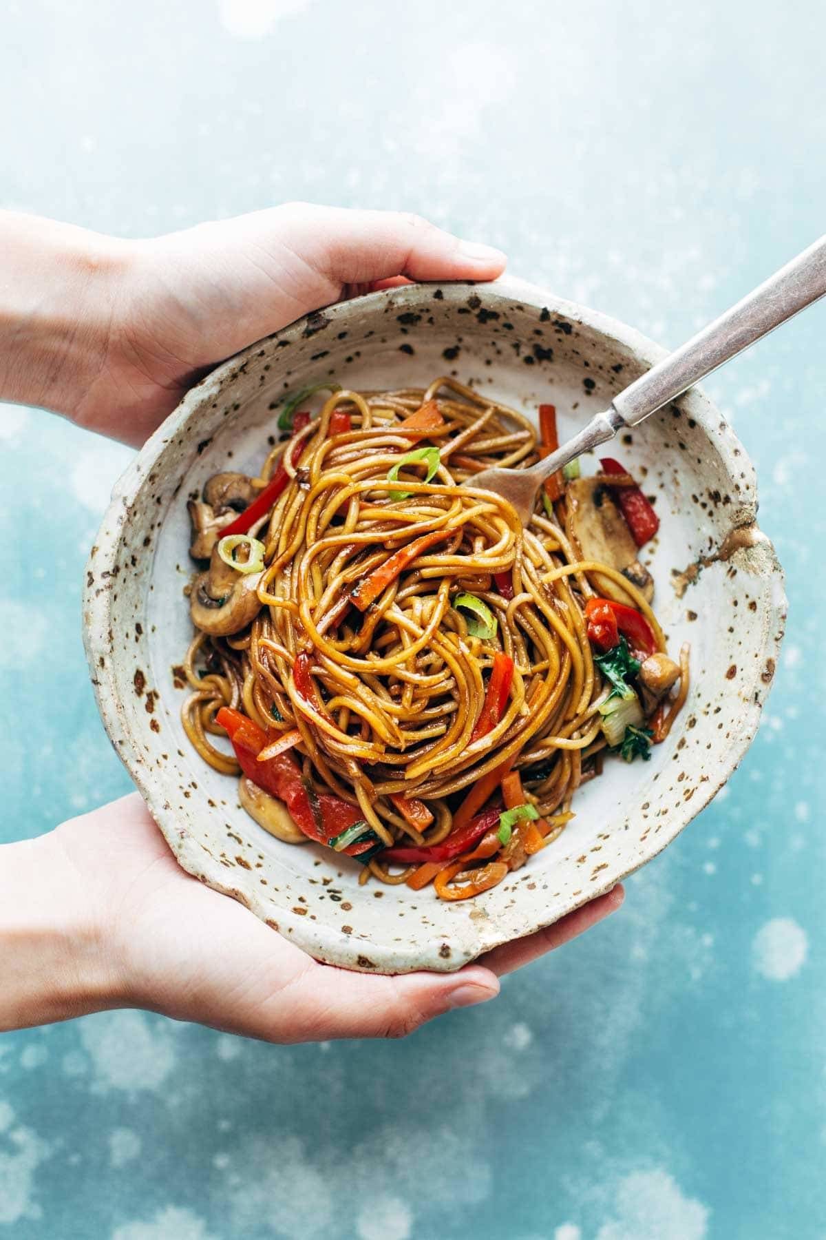 White hands holding a bowl of Lo Mein. Bowl has a fork in it.