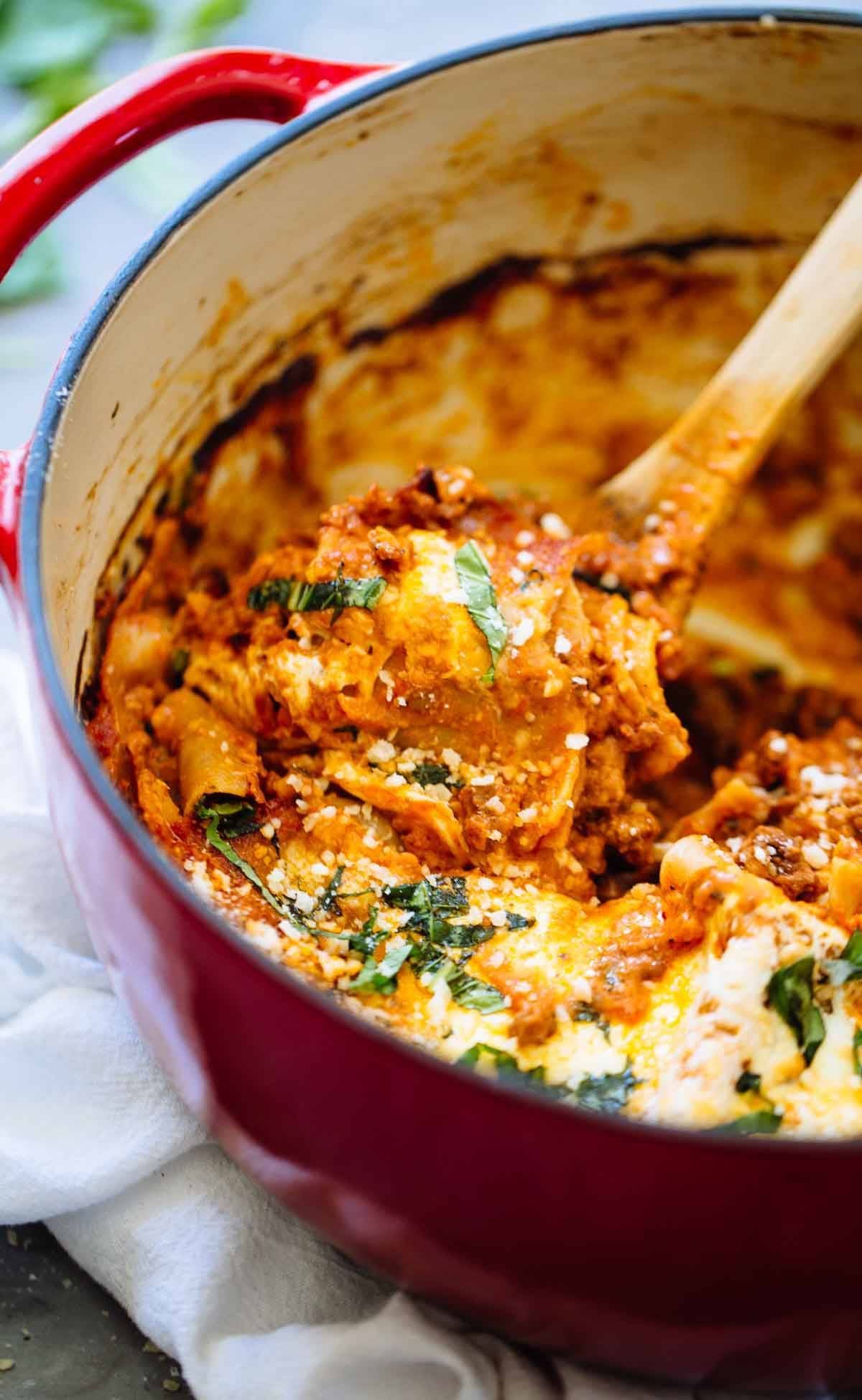 Lasagna in a red dutch oven with a wooden spoon.